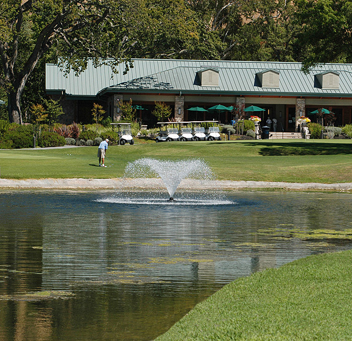 A golfer on the green behind a large water fountain and the club house in the background.